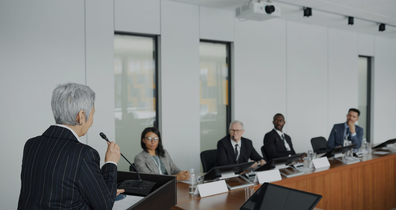 Senior executive presenting to board members in modern conference room with microphone and tablet.