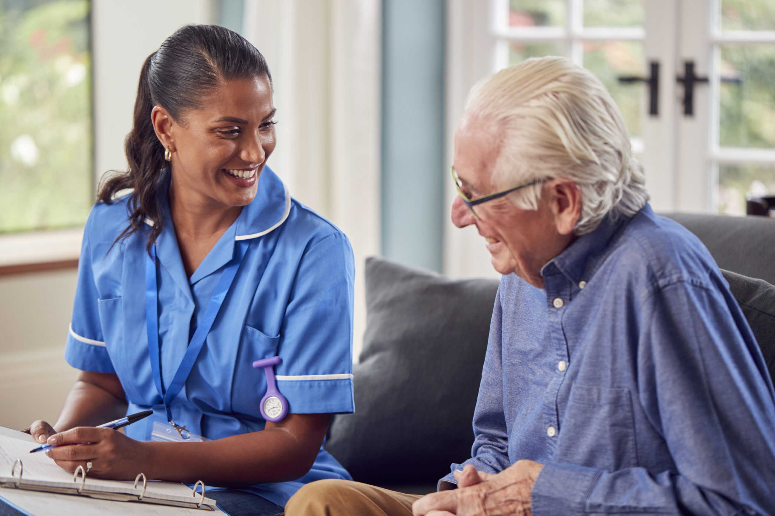 Kami Vision - Senior Man At Home Talking To Female Nurse Or Care Worker In Uniform Making Notes In Folder