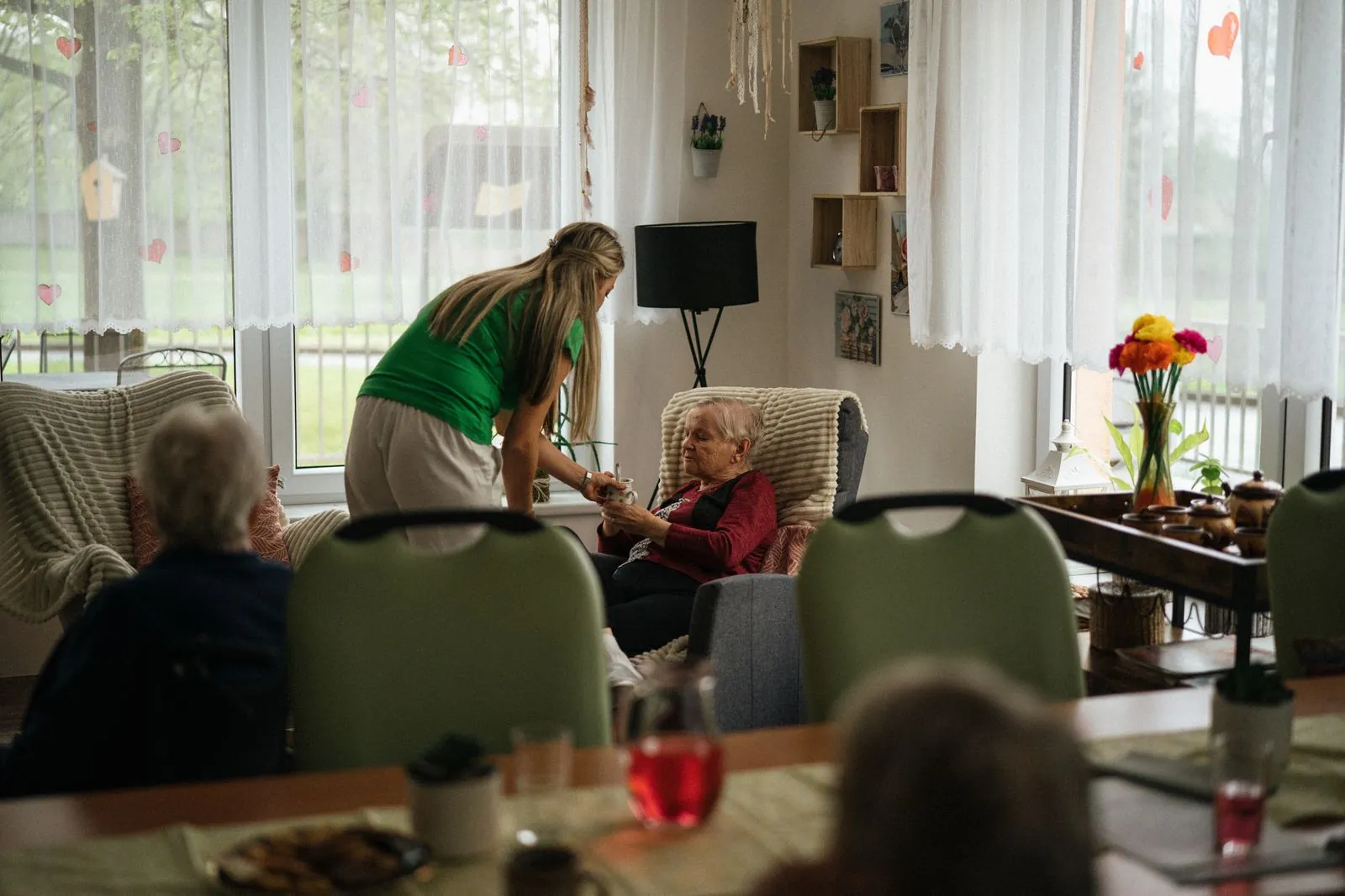A carer handing a cup of tea to an older woman in a bright communal lounge.
